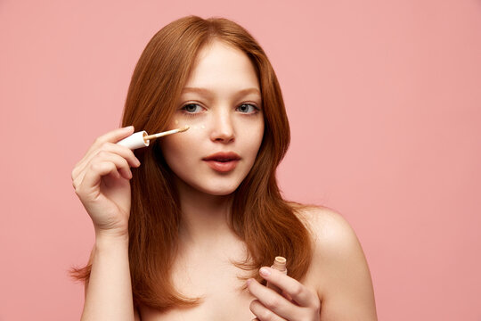 Portrait of young, beautiful, redhead girl with freckled skin applying concealer on face against pink studio background. Concept of natural beauty, makeup, cosmetology, cosmetics, skin care