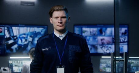 Male security officer in police monitoring center looks at camera. Multi ethnic employees work on computers on background. Big digital screens with surveillance CCTV cameras video footage. Portrait.