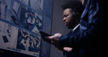 Security officer controls CCTV cameras using tablet and talks with African American coworker in...