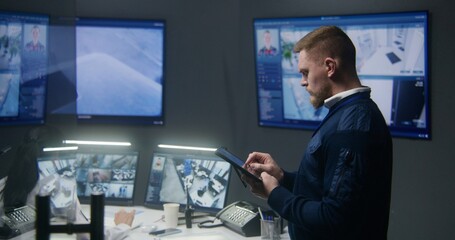Male officer controls security cameras in police surveillance center, uses tablet. PC monitors and multiple big screens on the wall with CCTV cameras footage. Security operators working at background.