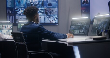 African American security operator monitors CCTV cameras with AI face recognition. Multiple big screens on the wall showing security cameras view. Colleague works at background. Social safety concept.