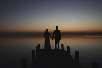 Groom and bride standing on wooden stage holding hands at sunset