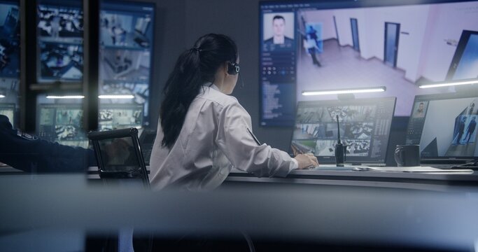 Female operator types keyboard and monitors security cameras with AI facial recognition in police surveillance room. PC monitors and big digital screens on the wall with displayed CCTV cameras view.