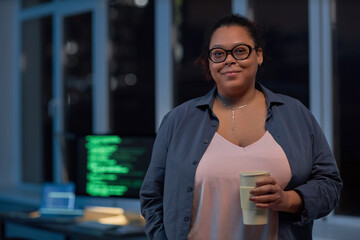 Portrait of African American woman in eyeglasses looking at camera while standing with coffee in IT office