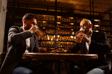 adult friends sitting with beer mugs in a pub. Two cheerful guys drink draft beer, celebrate meeting and talk