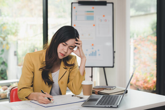 Young asian woman holding forehead while working on project at office desk. Tired stressful and upset businesswoman with graph report problem and question from project work.