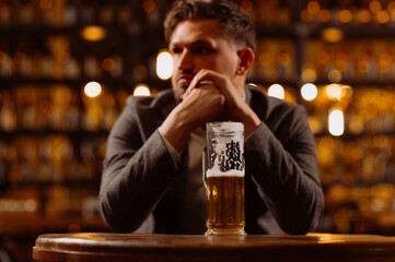 Sad young man in a suit looking at a glass of beer while sitting at a table in a pub. A man drinks after work in a pub.