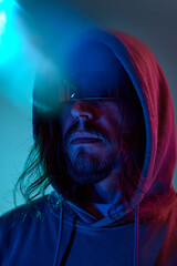 Portrait of serious man in hoodie and sunglasses against blue studio background in neon light. Night life. Futurism. Concept of human emotions, facial expression, lifestyle