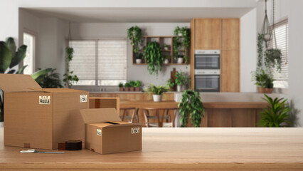 Wooden table, desk or shelf with stack of cardboard boxes over blurred view of kitchen and dining room, houseplants interior design, moving house concept with copy space