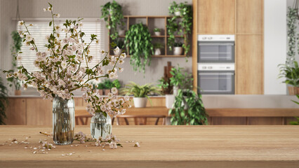 Wooden table, desk or shelf close up with branches of cherry blossoms in glass vase over blurred view of modern kitchen, urban jungle interior design concept