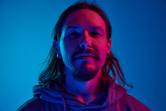 Portrait of man with moustache in casual clothes, hoodie looking at camera, posing with smile against blue studio background in neon light. Concept of human emotions, facial expression, lifestyle