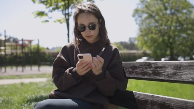 A Caucasian girl in dark sunglasses and a brown hoodie sits on a bench, an electronic cigarette in th park and holds a smartphone in her hand. The girl smokes e-cigarette and watching at smartphone.