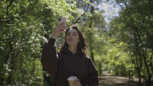 Сute caucasian girl photographs a beautiful landscape in a sunny park on a smartphone. Girl walks in the park and shoots on a mobile phone. The beautiful caucasian girl takes pictures of nature.