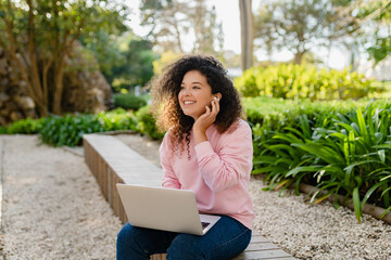 pretty curly young woman sitting in park working on laptop remote job