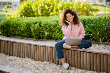 pretty curly young woman sitting in park working on laptop remote job