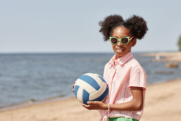 Portrait of African American little girl in sunglasses smiling at camera while playing volleyball on the beach
