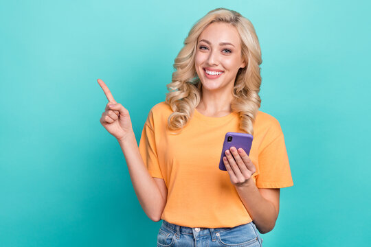 Photo of cheerful girl with curly hairstyle dressed yellow t-shirt directing empty space hold smartphone isolated on teal color background