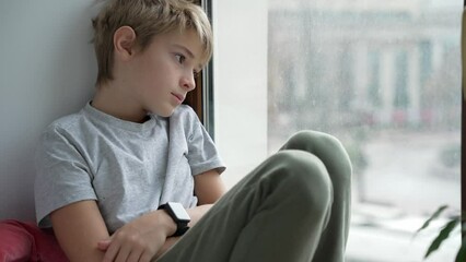 Boy is looking out of the window. Teenager sitting next to window at home alone and is sad or thinking about something. Emotional concept