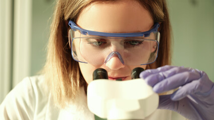 Woman scientist chemist in protective glasses looking through microscope in chemical lab. Laboratory diagnosis of internal diseases concept