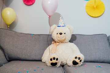 Big Teddy bear with balloons in party cap sitting on the gray sofa in decorated room. Birthday or festive holiday background. Selective focus, copy space