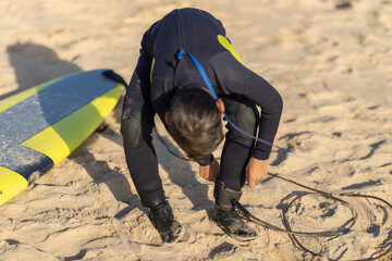 A man surfer fastens a board to his leg while sitting on the shore