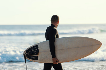 A surfer in a wetsuit walking on the shore holding a surfboard and looking at the sea