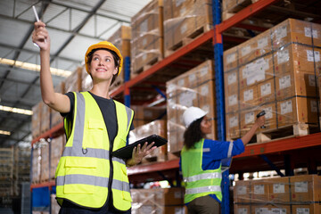 Woman intern working inside warehouse checking stock on shelves using clipboard - obrazy, fototapety, plakaty