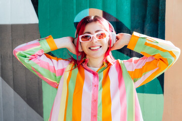 Emotional stylish young woman with pink hair and sun glasses in multicolor strippled shirt wears...