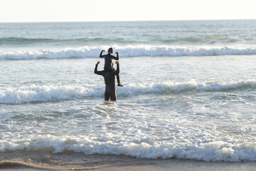 Little son on the shoulders of his father in the sea showing muscles - back biew