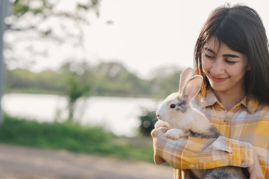 Portrait of woman embrass rabbit smile with happiness while rabbit stand with two front leg on woman arm with blurr nature green grass and pond background.