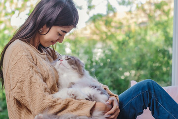 Woman with long hair in brown sweater and jean holding persain cat in her lap and embrassed him...
