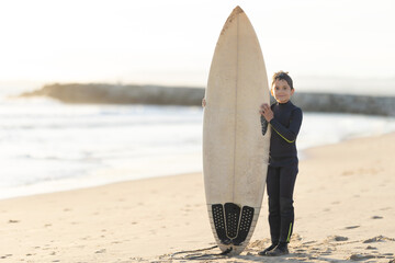 A little boy in a wetsuit holding a big white surfboard standing on the seashore