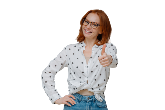 Pleasant looking confident woman with ginger hair, shows thumb up, says very good and encourages friend, likes someones idea, gives positive reply, wears stylish outfit, stands indoor over grey wall