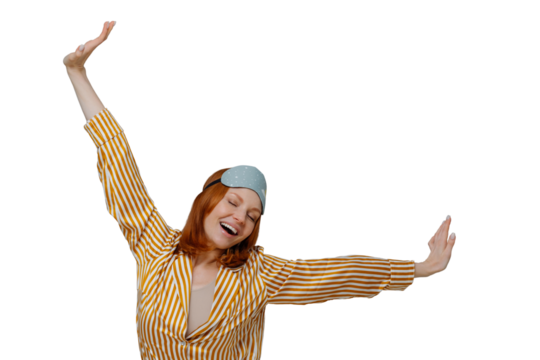Good morning and awakening concept. Happy relaxed red haired young woman wears comfortable soft pajamas and sleepmask, stretches arms sideways, has glad expression, isolated on grey background.