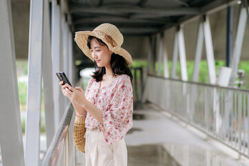 young asian woman traveler with weaving basket using mobile phone and standing on overpass. Journey trip lifestyle, world travel explorer or Asia summer tourism concept.