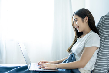 Fototapeta na wymiar Young smiling Asian woman in casual pajamas outfit sitting in bed in morning working on laptop, freelancer at home.