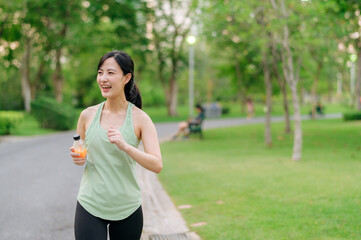 Female jogger. Fit young Asian woman with green sportswear drinking organic orange juice after running and enjoying a healthy outdoor. Fitness runner girl in public park. Wellness being concept