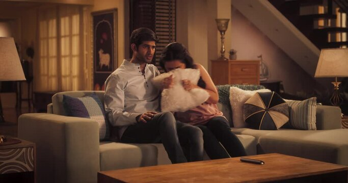 
Young Indian Couple Watching Scary Movie on TV, Finds Comfort In Each Other's Arms As They Sit On Their Couch. Boyfriend Holds His Dear Girlfriend and Supports Her. Horror Movie or News Concept