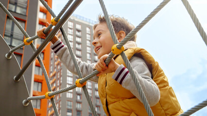 Cute smiling boy climbing up the rope net on the public playground at park. Active child, sports...