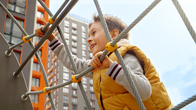 Cute smiling boy climbing up the rope net on the public playground at park. Active child, sports and development, kids playing outdoors - Powered by Adobe