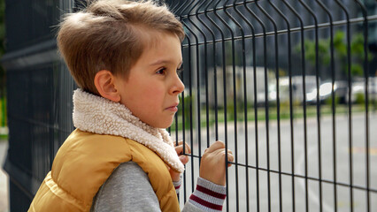 Portrait of lonely boy holding hands on metal fence and looking through it. Child depression,...