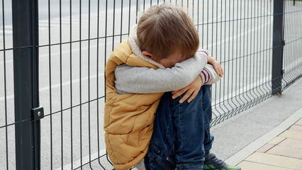 Lonely boy feeling alone on playground sitting next to metal fence and crying. Child depression,...