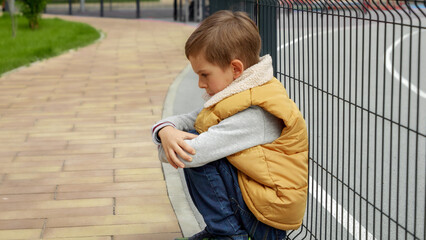 Unhappy lonely boy leaning on metal fence at school playground and sitting down. Child depression,...