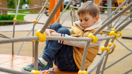 Upset lonely boy sitting on the playground and embracing his legs. Child depression, problems with...