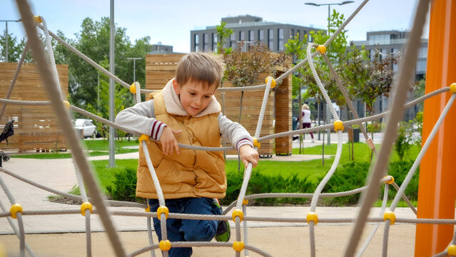 Cheerful boy climbing through ropes and obstacles on playground at modern living block outdoors