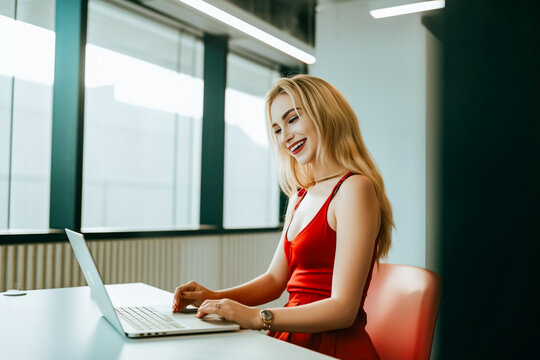 An alluring and fashionable blond lady in a stunning red dress focuses intently on her laptop while working amidst the picturesque outdoor field. generative AI.