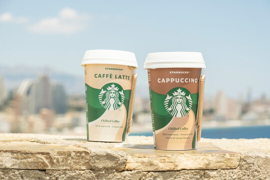 Starbucks coffee cup outdoors. Starbucks Cappuccino Chiled Coffee and Caffe Latte Chilled Coffee to go coffee cup with Benidorm city and Mediterranean Sea background. Benidorm, Spain - 15 May 2023