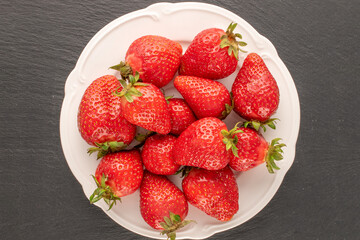 Several red sweet strawberries with white ceramic dish on slate stone, macro, top view.