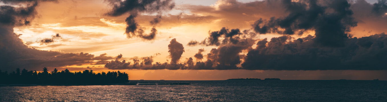 A stunning panoramic view of the sea during a breathtaking Maldivian sunset with orange sun behind dark-colored clouds and a silhouette of the trees in the land on the left side of the picture