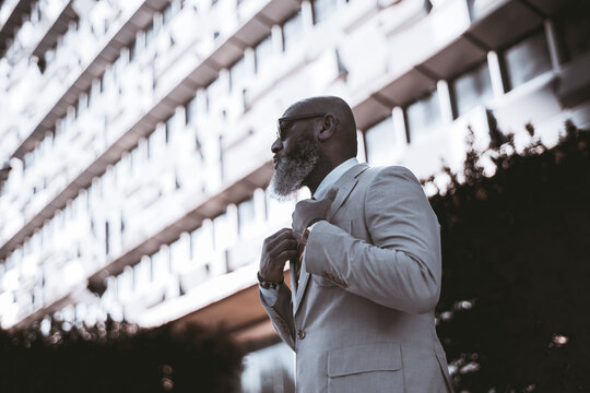 Lisbon; copy space; low angle shot of a bald man with a full beard showcasing white colored hair, wearing a stylish suit, adjusting his jacket, with a building defocused in the background